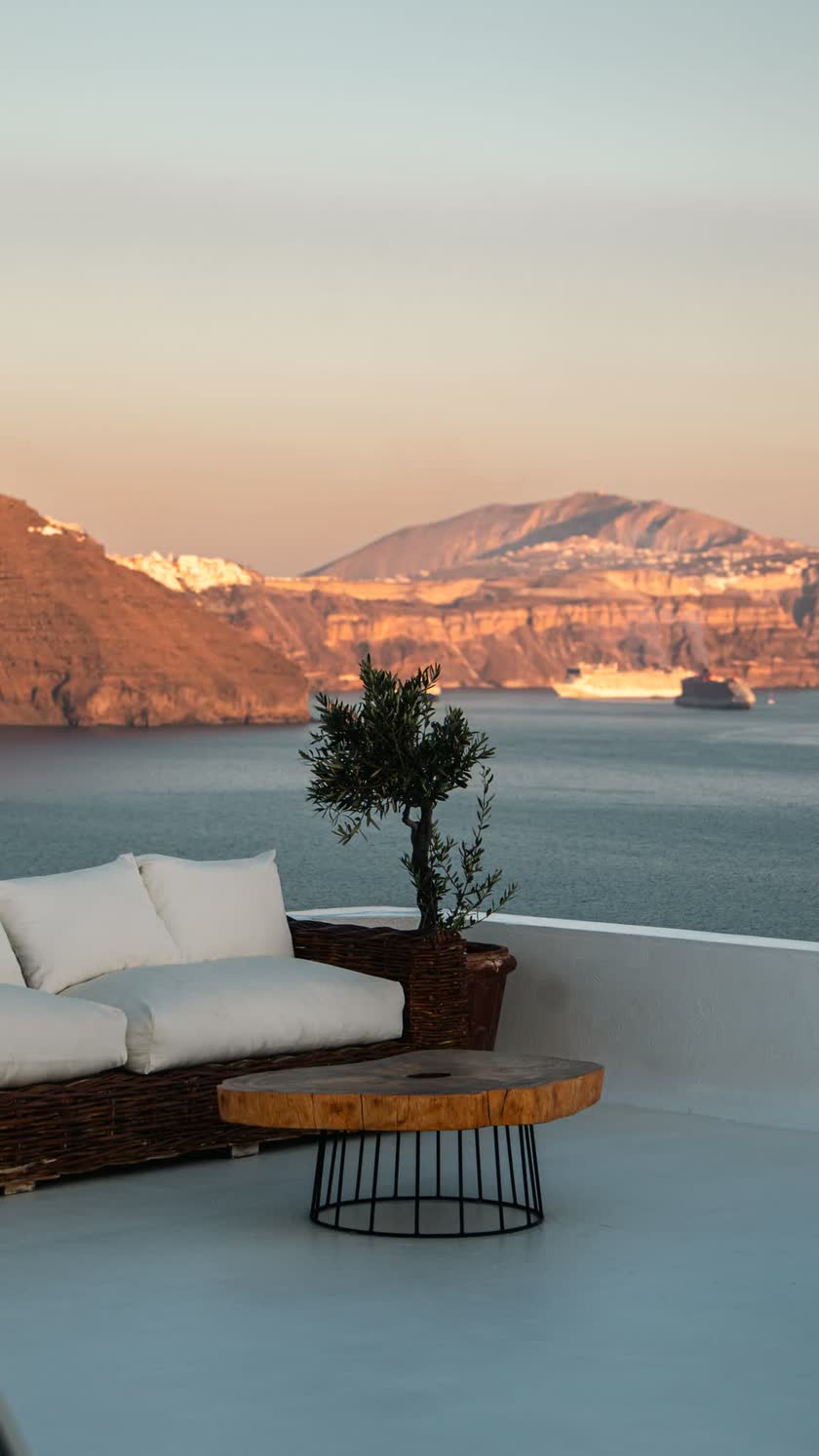 A listing photograph of a terrace at dusk, a wicker sofa and an olive tree, the sea and a caldera behind.